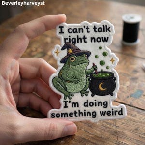 Può includere: Una toppa ricamata su sfondo bianco raffigurante una rana verde con un cappello da strega viola, intenta a mescolare un calderone nero con una luna crescente gialla. Il testo recita, I can't talk right now, I'm doing something weird.