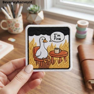 Puede incluir: Un parche bordado cuadrado que muestra un ganso blanco sentado en una silla junto a una mesa pequeña con una taza de café, rodeado de llamas amarillas y naranjas, una nube negra arriba y un globo de texto con la frase I'm Fine.