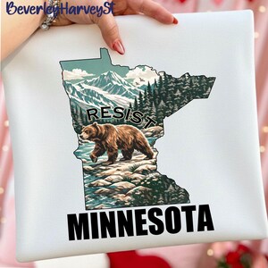 May include: White square pillow featuring a detailed illustration of the state of Minnesota. The design includes a bear, mountains, and trees, with the word "RESIST" above the bear and "MINNESOTA" below.