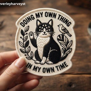 May include: A circular embroidered patch with a cream background and black stitching. It features a black and white cat with a multi-eyed face, a small bird, and leafy branches. The text reads DOING MY OWN THING IN MY OWN TIME.