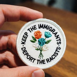 May include: A circular embroidered patch with a white background and a white stitched border. The text "KEEP THE IMMIGRANTS DEPORT THE RACISTS" is embroidered in black thread around the center, which features a bouquet of orange, teal, and white flowers.