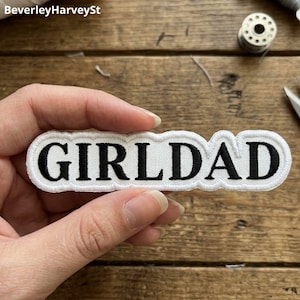 May include: A white embroidered patch with the text "GIRLDAD" in black, featuring a white border. The patch is held in a hand against a wooden background. The patch is rectangular in shape.
