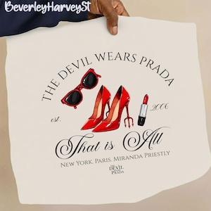 May include: Cream-colored tote bag featuring the text "THE DEVIL WEARS PRADA" with red sunglasses, red high heels, and a lipstick illustration. The text "That is All" is also present, along with the year 2006.