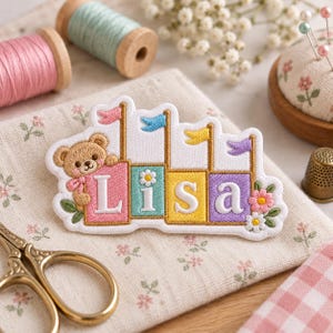 May include: An embroidered patch featuring a light brown teddy bear holding a pink block with the letter L, followed by blocks for i, s, and a in teal, yellow, and purple. Each block has a small flag on top, and the design includes flowers.