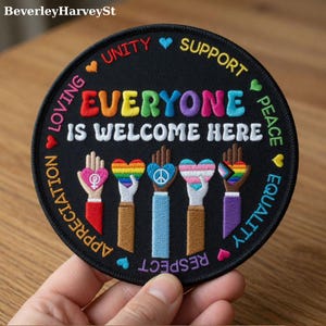 Op de afbeelding: Ronde zwarte geborduurde patch met de tekst "EVERYONE IS WELCOME HERE" in regenboogkleuren. De patch toont handen die harten vasthouden met verschillende trots symbolen, omringd door woorden als "UNITY" en "EQUALITY".