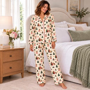 Puede incluir: Un conjunto de pijama de dos piezas que consta de una camisa de manga larga con botones y pantalones a juego en color crema. La tela presenta un patrón repetitivo de árboles de Navidad verdes, ángeles y escenas de nacimiento con ribetes rojos.