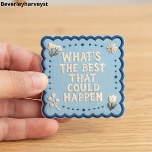 May include: A square embroidered patch with a scalloped dark blue border on a light blue background. The text "WHAT'S THE BEST THAT COULD HAPPEN" is stitched in white thread in the centre, surrounded by small embroidered white floral motifs.