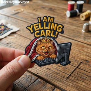 May include: An embroidered patch featuring an orange cat wearing a gold crown, sitting at a computer keyboard, and wrapped in a red blanket. The text above the cat reads I AM YELLING CARL! in bold yellow letters with a dark blue outline.