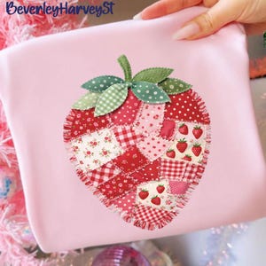 May include: A pink fabric item with a large, appliquéd strawberry design. The strawberry is made of various red and pink patterned fabrics, including polka dots, floral prints, and gingham. Green leaves and stem complete the design. The text "BeverleyHarveySt" is in the upper left corner.