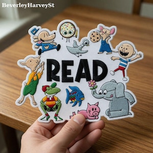 Puede incluir: Parche bordado con la palabra "READ" en letras negras, rodeado de personajes de dibujos animados coloridos. Los personajes incluyen un perro, un ratón, un cerdo y un elefante. El parche es blanco con un borde decorativo.