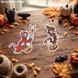 May include: Two embroidered patches featuring cartoon ducks in Halloween costumes. One duck wears a red devil suit with horns, a tail, and a pitchfork, holding a treat bag. The other duck wears a pointed orange and brown witch hat and cape.