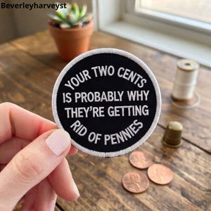 Könnte beinhalten: Ein kreisförmiger, schwarz bestickter Aufnäher mit weißem Rand und weißem Text, der lautet: YOUR TWO CENTS IS PROBABLY WHY THEY'RE GETTING RID OF PENNIES. Der Aufnäher hat eine saubere, gestickte Oberfläche und wird vor einer rustikalen Holzoberfläche gehalten.