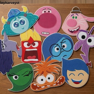 May include: A collection of nine embroidered character patches featuring various colors and facial expressions, including a teal monster, a pink circle, a light pink creature, a purple figure, a red block, a blue character with glasses, a green shape, an orange creature, and a blue face.