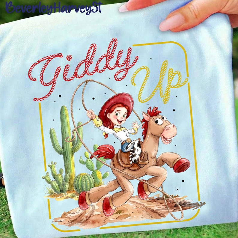 May include: Pale blue t-shirt with a cartoon illustration of a cowgirl riding a horse, and the words "Giddy Up" in red and yellow. The cowgirl wears a red hat and holds a lasso. Cacti are in the background.