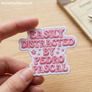 Könnte beinhalten: Ein weiß-rosa bestickter Aufnäher mit dem Text "EASILY DISTRACTED BY PEDRO PASCAL". Der Aufnäher hat einen weißen Rand und rosa Schrift. Der Aufnäher wird von den Fingern einer Person gehalten.