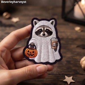 Pode incluir: Um emblema bordado apresentando um guaxinim vestindo uma fantasia de fantasma branca. O guaxinim segura um balde de abóbora laranja em uma mão e um copo de café bege com um ícone de caveira na outra. O emblema tem uma borda roxa escura.