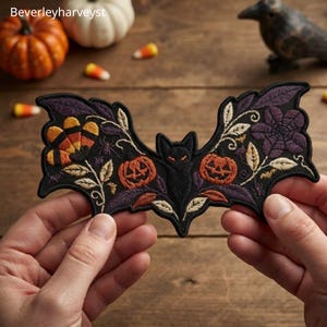 Puede incluir: Un parche bordado de murciélago negro con motivos de dulces de maíz naranjas y amarillos, dos calabazas de Halloween naranjas y detalles florales morados. El diseño incluye costuras detalladas de hojas y pequeñas arañas sobre tela.