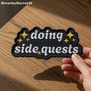 May include: Black embroidered patch with the words "doing side quests" in white script. The patch has a black border and yellow star accents. The patch is held by a hand.
