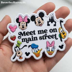 Op de afbeelding: Wit geborduurd embleem met Mickey Mouse, Minnie Mouse, Goofy, Donald Duck en Daisy Duck. De patch bevat de tekst "Meet me on main street" in een decoratief lettertype, plus een kasteel en een hart.