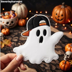 May include: An embroidered patch featuring a white ghost with a surprised expression, wearing a black baseball cap with an orange brim. The ghost has black oval eyes and a black mouth, with detailed white stitching throughout the fabric design.