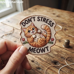May include: A circular embroidered patch featuring an orange tabby cat lying on its back with its tongue out. The text DON'T STRESS MEOWT is stitched in dark brown thread. Small pink flowers and green leaves are embroidered at the base of the cat.