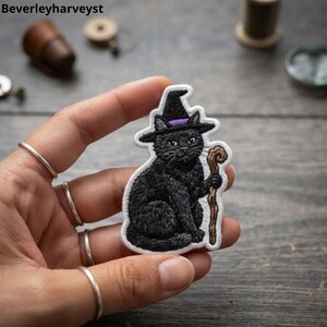May include: An embroidered patch featuring a black cat wearing a pointed black hat with a purple band. The cat is sitting and holding a brown wooden staff. The patch has a white border and is shaped to follow the outline of the cat and staff.