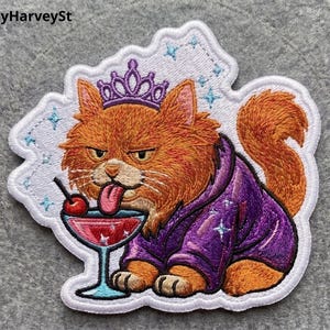 May include: An embroidered patch featuring an orange cat wearing a purple crown and a purple robe. The cat has its tongue out, licking a red cherry inside a cocktail glass. The design includes small blue star accents on a white background.