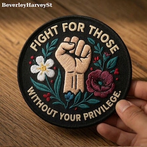 Può includere: Toppa rotonda ricamata nera con la scritta "FIGHT FOR THOSE WITHOUT YOUR PRIVILEGE". La toppa presenta un pugno chiuso, fiori e foglie in bianco, rosso e verde. La toppa è tenuta da una mano.