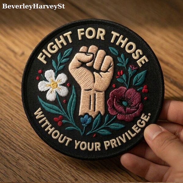 May include: Circular black embroidered patch with the text "FIGHT FOR THOSE WITHOUT YOUR PRIVILEGE." The patch features a raised fist, flowers, and leaves in white, red, and green. The patch is held by a hand.