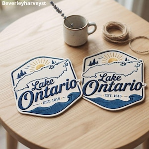 May include: Two hexagonal fabric coasters featuring an embroidered map of Lake Ontario in navy blue and cream. The design includes a yellow sun, pine trees, waves, and the text Lake Ontario EST. 1615. The coasters are placed on a light wood surface.