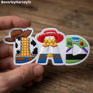 May include: Embroidered patch spelling "DAD" with letters designed as characters from the Toy Story film. The "D" is Woody, the "A" is Jessie, and the "D" is Buzz Lightyear. The patch is white with colourful details.