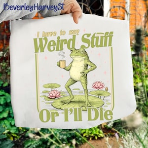 May include: White tote bag featuring a cartoon frog holding a coffee cup, standing on a lily pad. The text "I have to say Weird Stuff Or I'll Die" is printed in green. Pink lotus flowers and lily pads are also visible.