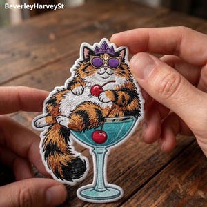 Op de afbeelding: Een geborduurde patch met een pluizige lapjeskat die een paarse kroon en paarse zonnebril draagt. De kat ligt in een lichtblauw cocktailglas en houdt een rode kers met steeltje vast. De patch heeft een witte rand en gedetailleerd stikwerk.