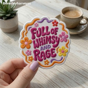 Puede incluir: Un parche bordado circular con fondo blanco que presenta el texto FULL OF WHIMSY AND RAGE en letras de color rosa oscuro. El diseño está decorado con flores naranjas, amarillas y rosas y líneas onduladas moradas, todo rodeado por un borde naranja.