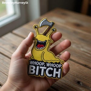 May include: A yellow embroidered patch featuring a cartoon slug with a wide, toothy grin and a red tongue, holding a grey axe with a brown handle. Below the slug, the text WHOOP, WHOOP BITCH is embroidered in bold white capital letters.