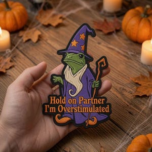 May include: An embroidered patch featuring a green frog dressed as a wizard in a purple robe and hat with orange stars. The frog holds a staff and wears orange curled shoes. The text reads: Hold on Partner I'm Overstimulated.