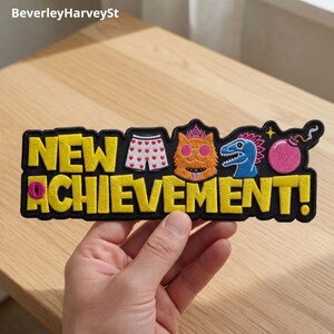 Op de afbeelding: Een zwart geborduurd embleem met de woorden "NEW ACHIEVEMENT!" in gele blokletters. Het embleem toont cartoonafbeeldingen van ondergoed, een kat, een dinosaurus en een bom.
