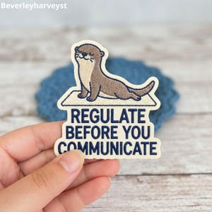 May include: An embroidered patch featuring a brown otter lying on a cream-colored mat. Below the otter, the text reads REGULATE BEFORE YOU COMMUNICATE in dark blue capital letters. The patch has a cream-colored border and a die-cut shape.
