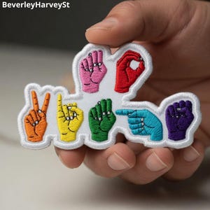 May include: An embroidered patch featuring seven hands in various American Sign Language positions, each in a different color including pink, red, orange, yellow, green, blue, and purple, arranged on a white background with a white border.