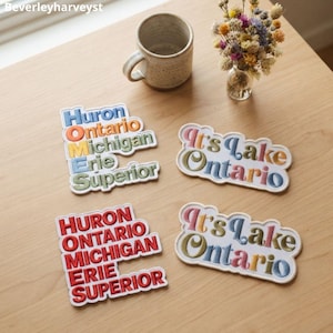 Op de afbeelding: Vier geborduurde patches met tekst. Twee patches vermelden de Grote Meren: Huron, Ontario, Michigan, Erie en Superior, één met meerkleurig garen en één in het rood. Twee patches bevatten de tekst It's Lake Ontario in meerkleurig garen.