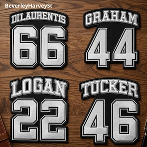 May include: Four black and white embroidered patches with names and numbers. The names are Dilaurentis, Graham, Logan, and Tucker. The numbers are 66, 44, 22, and 46. The patches are on a wooden surface.