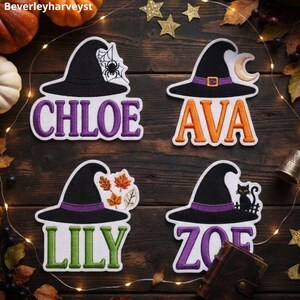 Pode incluir: Quatro emblemas bordados com chapéus de bruxa pretos e faixas roxas. Os emblemas exibem os nomes CHLOE em roxo, AVA em laranja, LILY em verde e ZOE em roxo. Os elementos decorativos incluem uma teia de aranha, uma lua crescente, folhas e um gato.