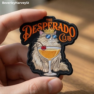 May include: An embroidered patch featuring a fluffy tan cat wearing a gold crown and blue sunglasses. The cat sits behind a cocktail glass with a red cherry. The text reads THE DESPERADO CLUB in orange letters against a black background.