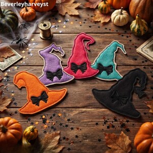 May include: Five felt witch hat cutouts in orange, purple, pink, teal, and black, each featuring a stitched black bow detail. The hats are arranged on a wooden surface surrounded by scattered autumn leaves, small pumpkins, and star-shaped confetti.