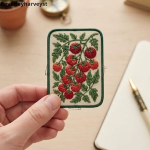 May include: A rectangular embroidered patch featuring a cluster of red tomatoes on green vines against a cream-colored background. The patch is bordered by a dark green stitched edge and displays detailed thread work of leaves and fruit.