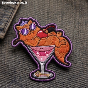 May include: An embroidered patch featuring an orange cat wearing purple sunglasses and a small crown, lounging inside a martini glass filled with a pink beverage and a red cherry. The design includes a gold key at the base of the glass.
