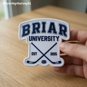 May include: A white embroidered patch featuring the words "Briar University" in blue. Below are crossed hockey sticks and a puck. The patch also includes the text "EST 2015".