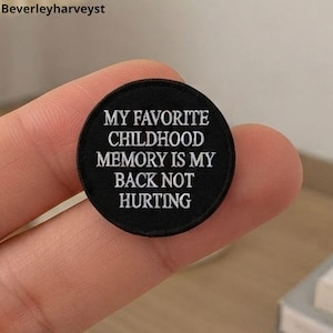 May include: A circular black embroidered patch featuring white text that reads, MY FAVORITE CHILDHOOD MEMORY IS MY BACK NOT HURTING. The patch has a clean, stitched border and is presented as a small, round fabric accessory with clear lettering.