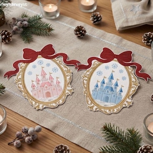 May include: Two oval-shaped decorative ornaments featuring a gold-colored frame and a red ribbon bow at the top. One ornament displays a pink castle with blue snowflakes, while the other shows a blue castle with blue snowflakes on a white background.