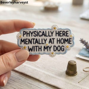 May include: An embroidered patch with a light blue scalloped border on a white background. The text reads PHYSICALLY HERE MENTALLY AT HOME WITH MY DOG in black thread. Small white and yellow daisy flowers are stitched around the border.